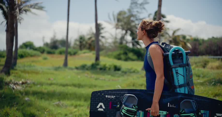 A determined female kitesurfer smiles as she walks along a lush tropical path, carrying her board and gear for a day on the water