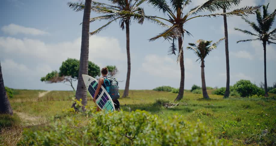 A female surfer with a backpack carries her surfboard as she walks through a grassy field with palm trees towards the beach