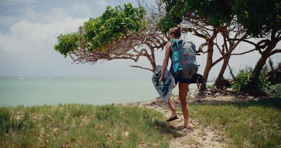 Female kitesurfer carrying her board and gear walks along a sandy path to a beautiful tropical beach