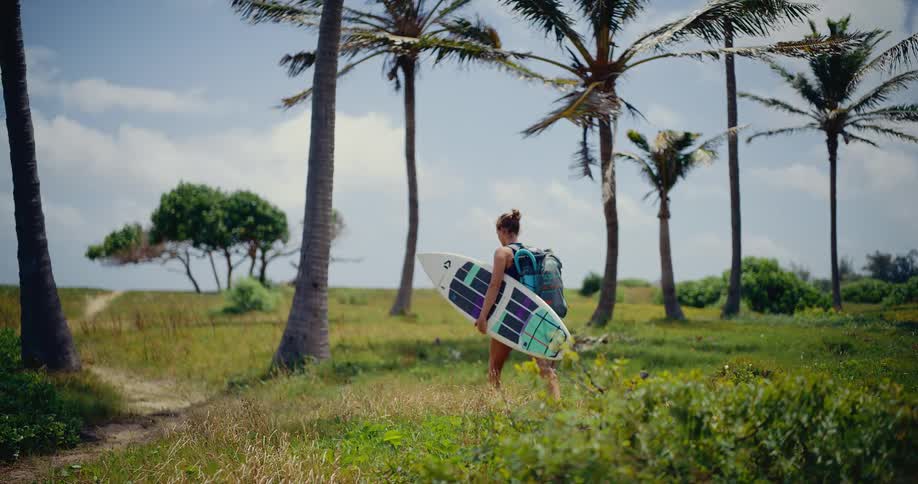 An adventurous woman carries her surfboard and backpack while walking through a tropical palm tree grove towards the beach in slow motion