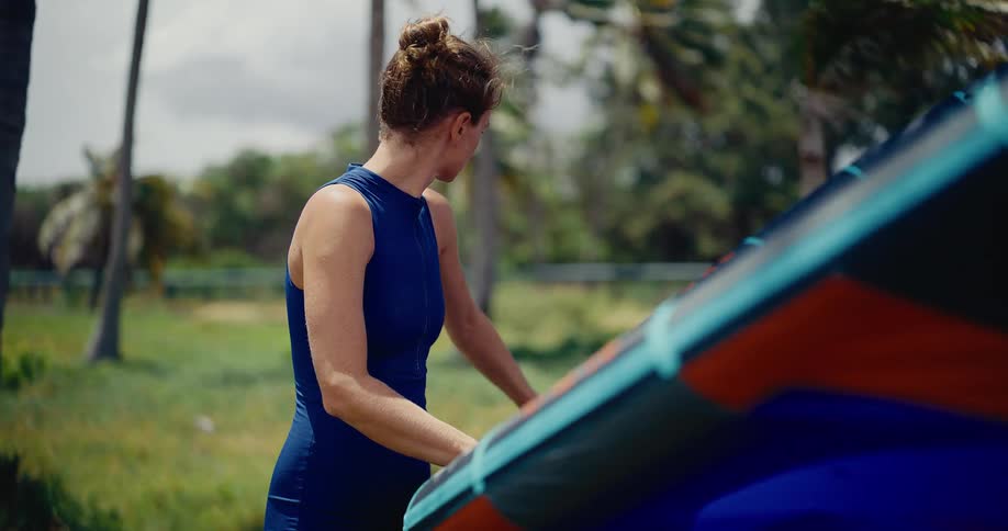A female kitesurfer in a blue wetsuit meticulously prepares her colorful kite on a windy, tropical day surrounded by palm trees