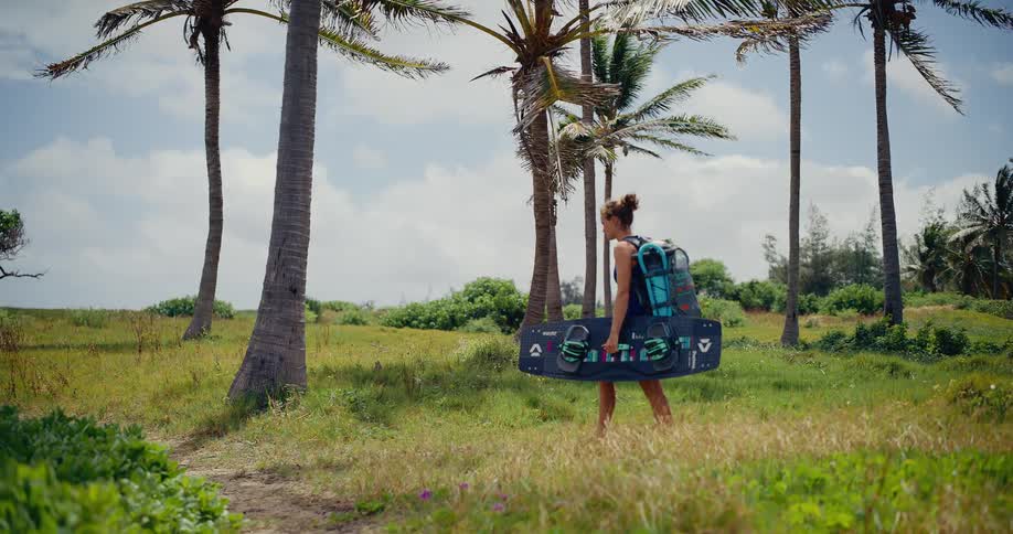 An athletic young woman carries her kiteboard and gear as she walks along a grassy path through a grove of palm trees on a windy tropical day