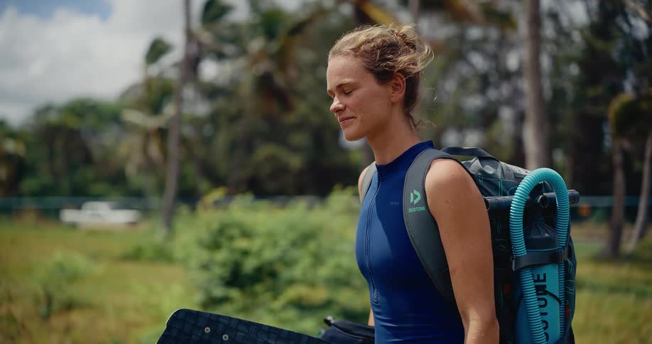 A happy female kitesurfer smiles as she walks with her equipment through a tropical landscape, ready for an adventure on the water