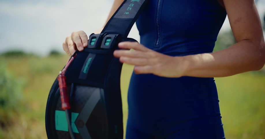 Close-up slow-motion shot of a female kitesurfer in a wetsuit putting on her harness in a grassy field
