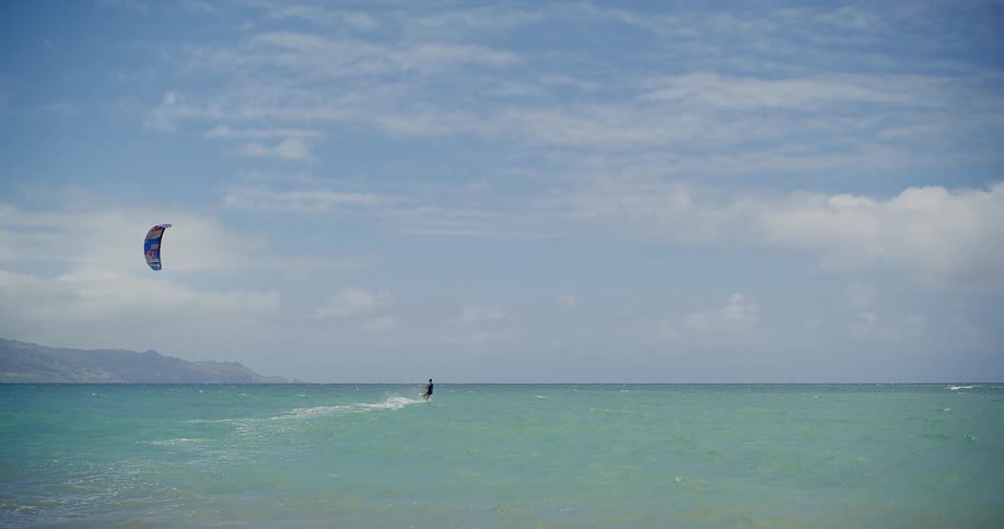 A lone kitesurfer glides across the vast turquoise ocean under a bright, cloudy sky
