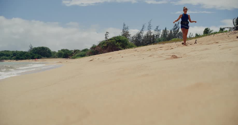 A female kitesurfer in slow motion walks along a sandy tropical beach carrying her kite lines and control bar