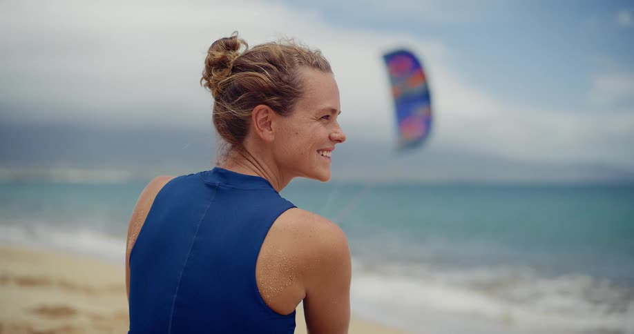 A smiling female kitesurfer sits on a sandy beach looking out at the ocean with a kite visible in the background