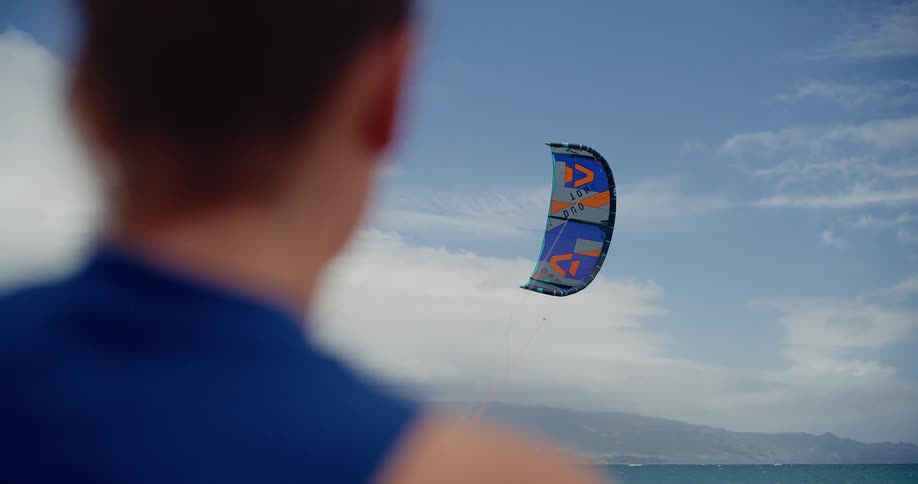A colorful kitesurfing kite floats in the sky, viewed from over the shoulder of a person on the beach