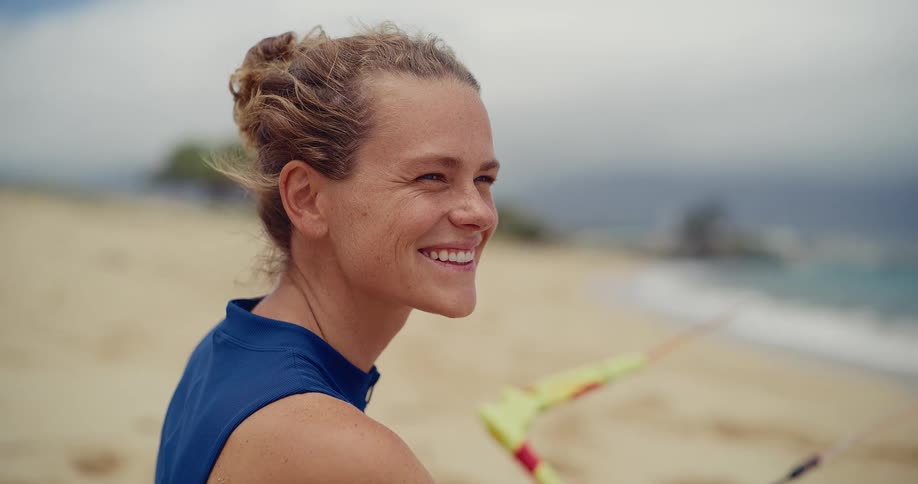 A close-up portrait of a happy, athletic woman with her hair in a bun sitting on a sandy beach and smiling as she looks out at the ocean