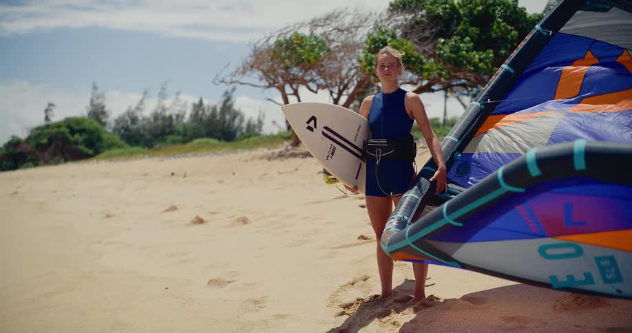 A young athletic woman in a wetsuit stands on a sandy beach, holding her kitesurfing board and kite, smiling confidently at the camera on a sunny, windy day