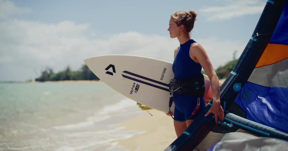A female kitesurfer in a wetsuit holds her board and kite while standing on a tropical beach, looking out at the ocean