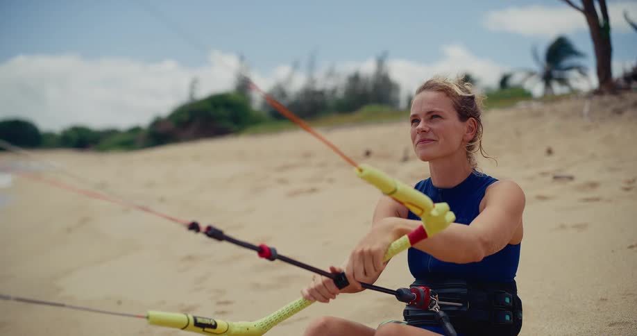 A beautiful young female kitesurfer smiles while sitting on a sandy beach, holding her kite bar and looking out at the windy ocean conditions