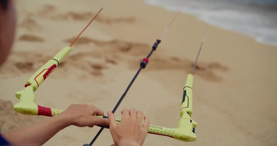 Over-the-shoulder close-up of a kitesurfer adjusting the control bar on a sandy beach before going into the water