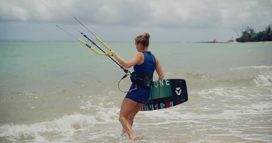 A female kitesurfer in a blue wetsuit walks from the sandy beach into the ocean waves in slow motion, ready to start her session