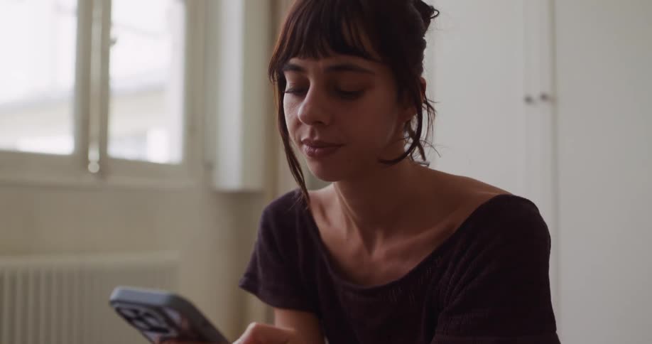 A young woman smiles softly while scrolling through her smartphone in a naturally lit room