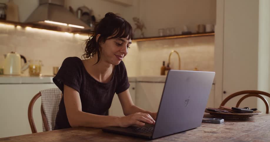 A young woman with dark hair sits at a wooden table in her modern kitchen, smiling as she types on her laptop