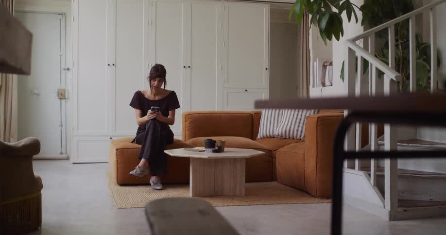 A young woman smiles while scrolling on her smartphone, relaxing on the couch in her modern, sunlit living room