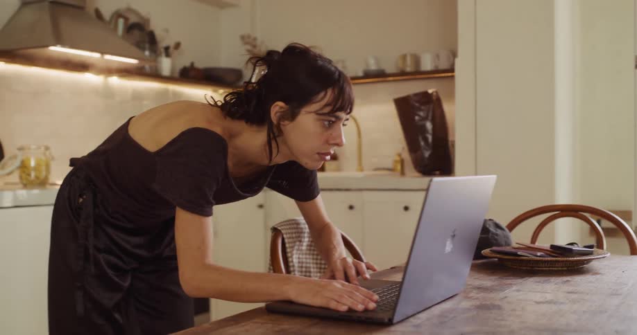 A focused young woman walks from her stove to her laptop to continue working while her dinner cooks in the background