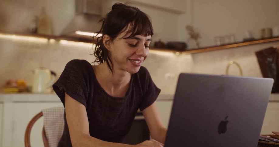 A smiling young woman happily types on her laptop while sitting at a wooden table in her kitchen