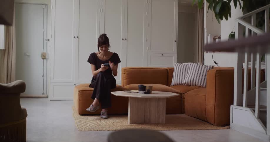 A thoughtful woman relaxes on a cozy brown sofa in her bright, modern living room and smiles while using her smartphone