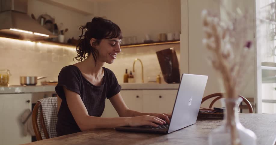 A happy young woman smiles while typing on her laptop at a rustic wooden table in a warm and cozy home kitchen
