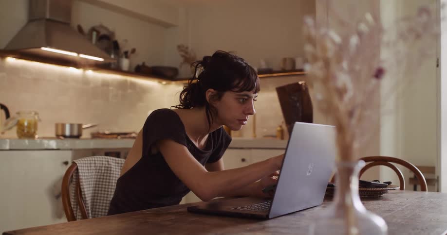 A focused woman works from home at her kitchen table, multitasking between her laptop and smartphone