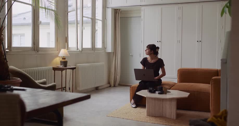 A pensive woman works from home on her laptop, pausing to look thoughtfully out the window of her stylish apartment