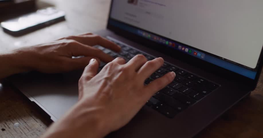 A close up of a person's hands as they type on a laptop keyboard while sitting at a wooden desk
