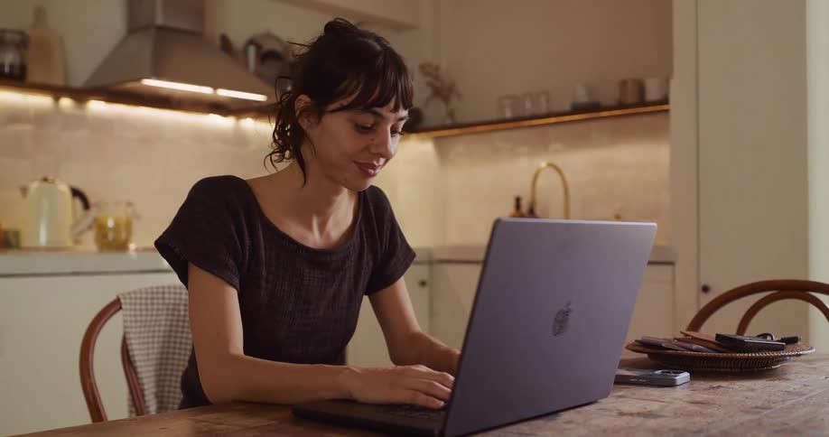 A smiling woman types on her laptop while sitting at a wooden table in her cozy kitchen