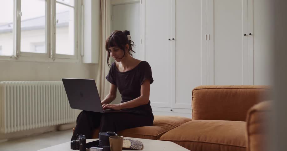 A female photographer works from home, typing on her laptop while sitting on the couch in her bright, modern apartment
