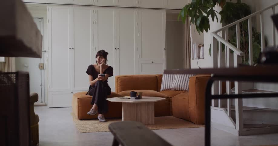 A woman relaxes on a comfortable orange couch in her modern living room, pausing from her phone to smile and daydream