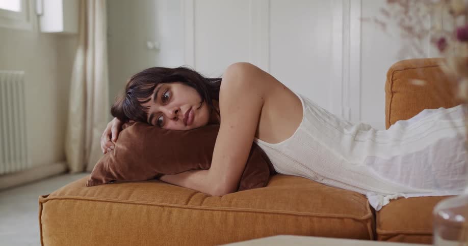 A pensive young woman with a sad expression lies on an orange couch in her sunlit living room, lost in thought