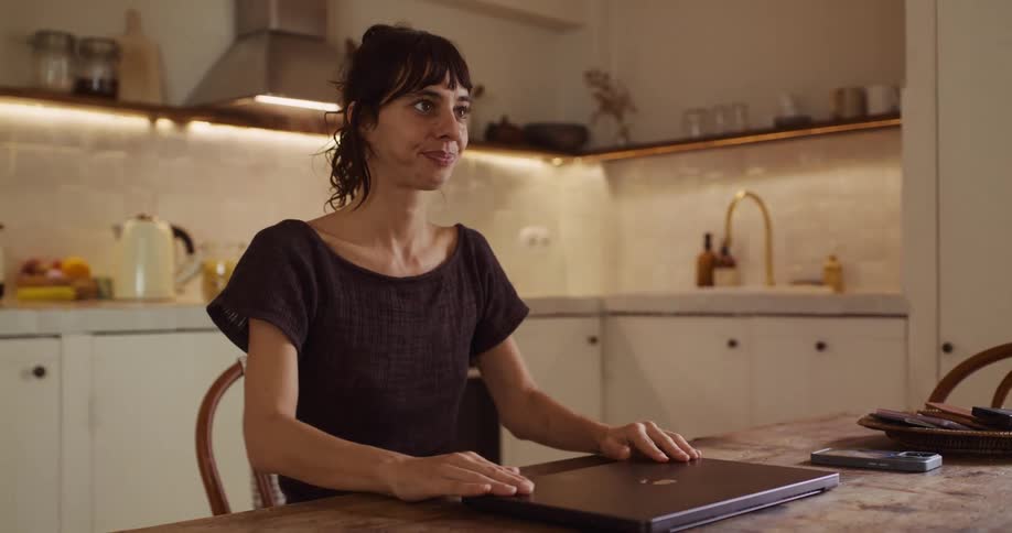A woman finishes her work on a laptop, closes it with a smile, and begins cooking dinner in her cozy kitchen