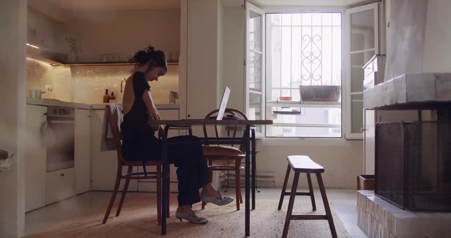 A young woman stretches her neck while working from home on her laptop in her sunlit apartment