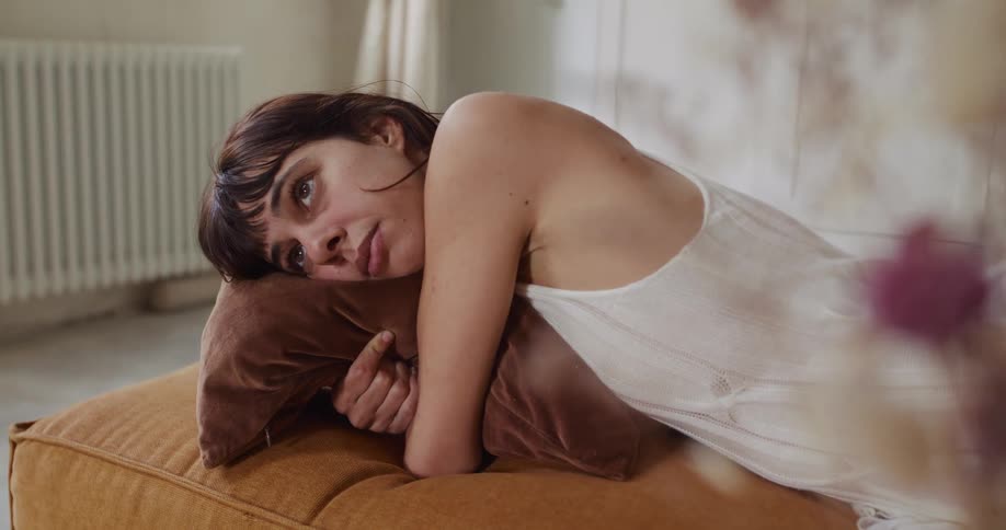 A thoughtful young woman wearing a white top rests peacefully on a floor cushion, cuddling a brown pillow in her sunlit apartment