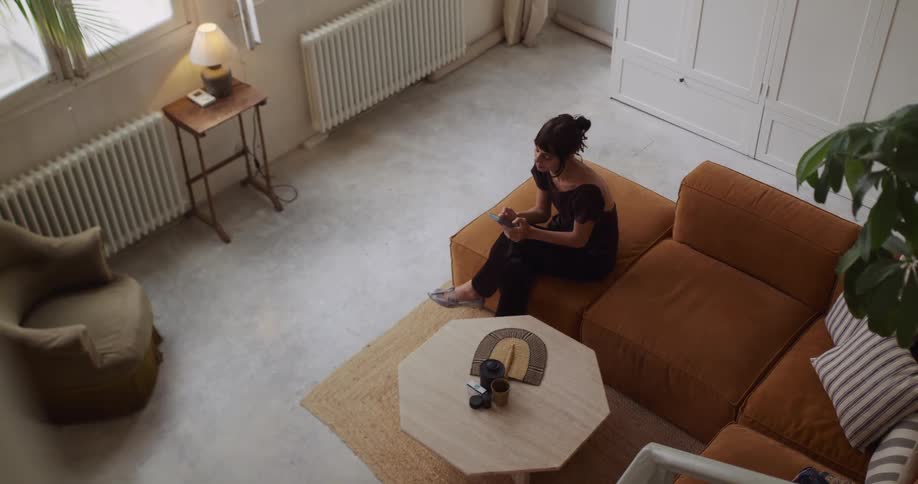 High-angle view of a woman relaxing on a stylish brown sofa in her modern loft, casually browsing her smartphone