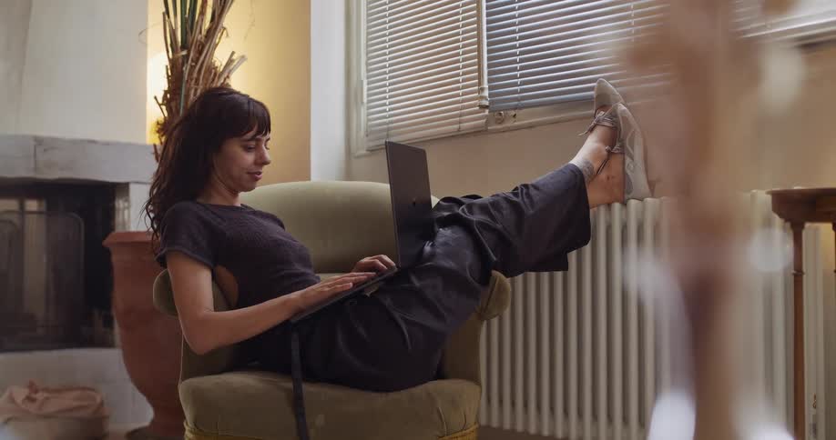 A woman casually works on her laptop, relaxing in an armchair with her feet propped up on the windowsill