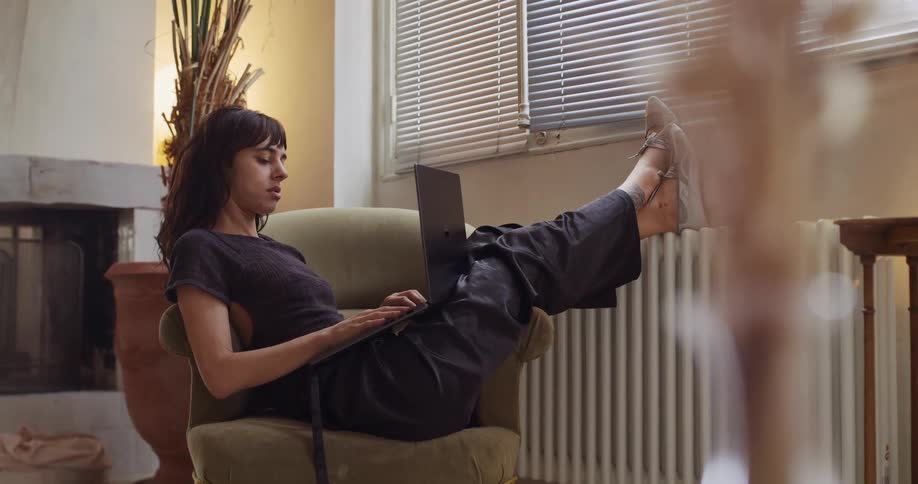 A relaxed woman sits in a comfortable armchair with her feet up, working on her laptop in a cozy living room