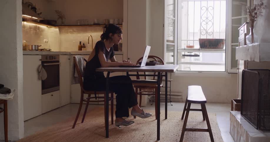 A stylish woman works on her laptop at a dining table in her sunlit apartment with the kitchen visible in the background