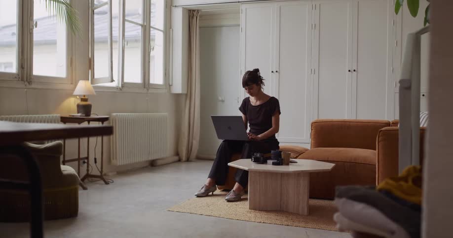 A focused woman works on her laptop in a bright and modern living room, pausing thoughtfully while looking out the window