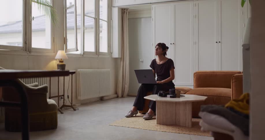 A creative woman works on her laptop, pausing to think, in a bright and spacious modern apartment