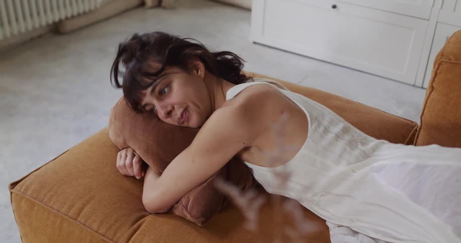 A melancholic young woman in a white dress slowly wakes from a nap on a mustard-colored couch in a minimalist apartment