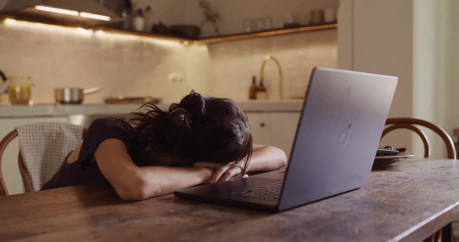 An overworked woman stares intently at her laptop screen before finally giving in to exhaustion and resting her head on the kitchen table