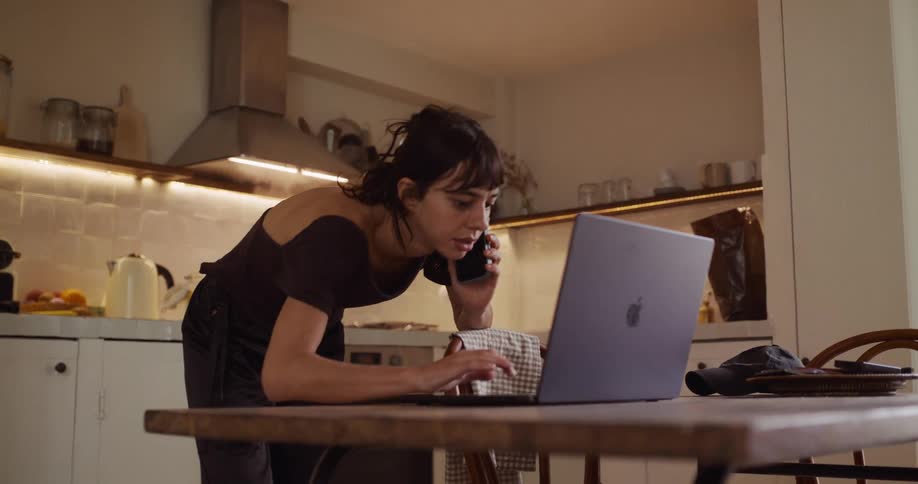 A busy young woman multitasks in her kitchen, stirring a pot on the stove while talking on the phone and then checking her laptop