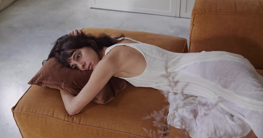 A contemplative woman in a white dress rests on a modern ochre sofa, with gentle sunlight and plant shadows drifting over her