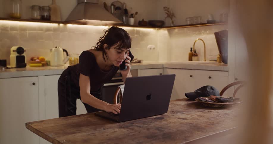 A busy woman multitasks in her cozy kitchen, talking on the phone and cooking while working on her laptop