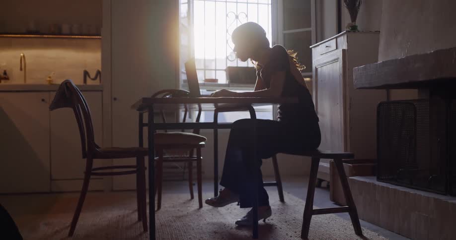 A focused woman works on her laptop at a rustic dining table, silhouetted against a bright, sunlit window