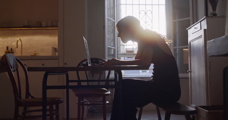 A woman is silhouetted by the bright sun streaming through a window as she works on her laptop at a dining table in her home