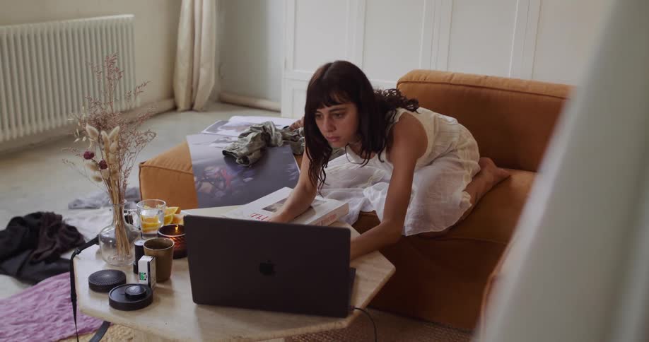 A focused woman in a white dress works from home on her laptop in a messy, sunlit living room