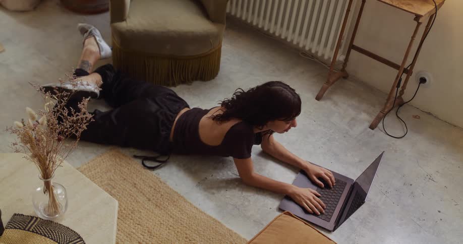 A relaxed woman lies on the floor of her apartment, comfortably working and browsing on her laptop